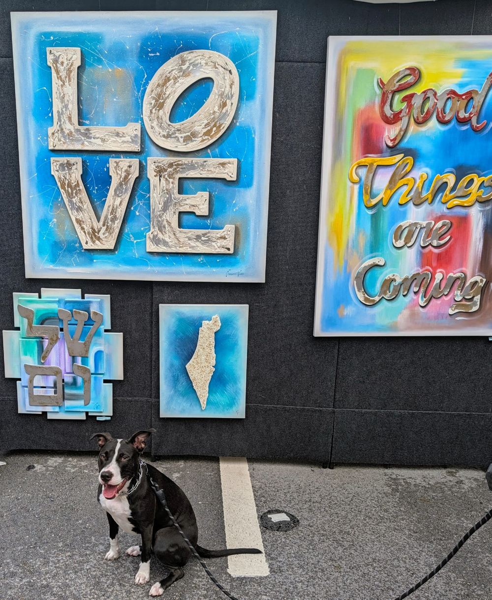 Bamba, a black and white dog, sitting in front of colorful artwork including a LOVE painting, a Shalom piece, and a map of Israel at the Las Olas Art Fair